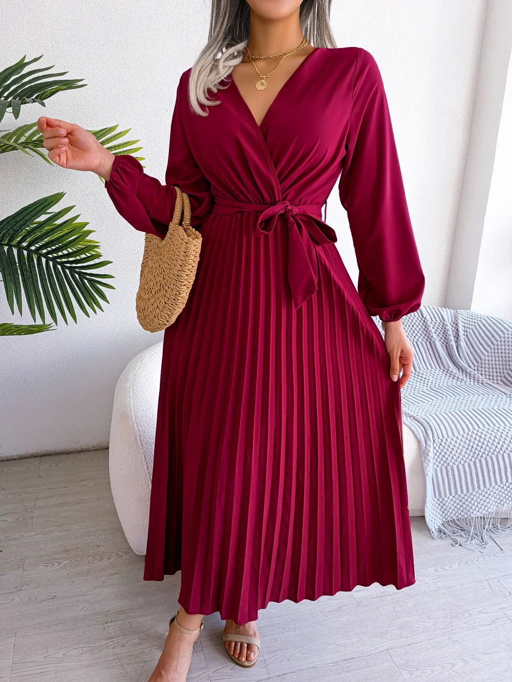 Woman wearing a burgundy pleated dress holding a leaf, standing in a room with a white couch and plant.