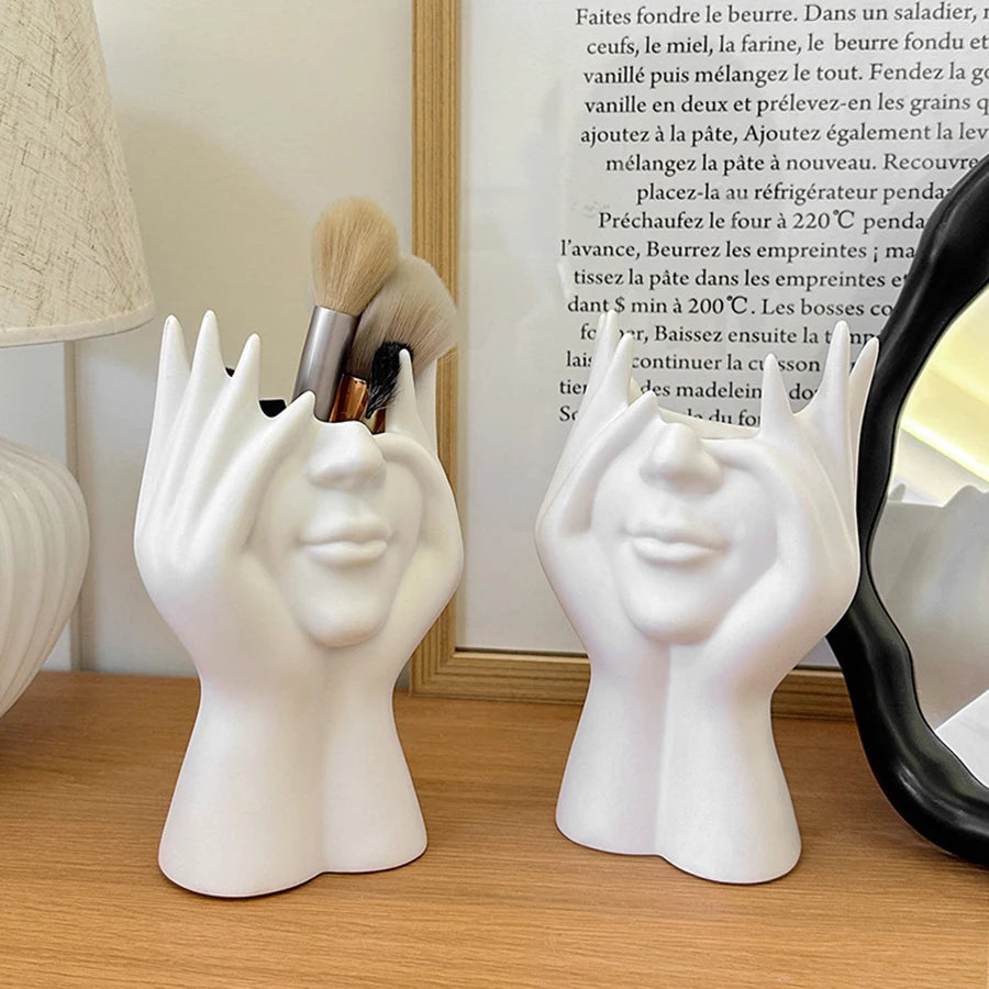 White ceramic face-shaped holders on a wooden surface with a mirror and lamp in the background.
