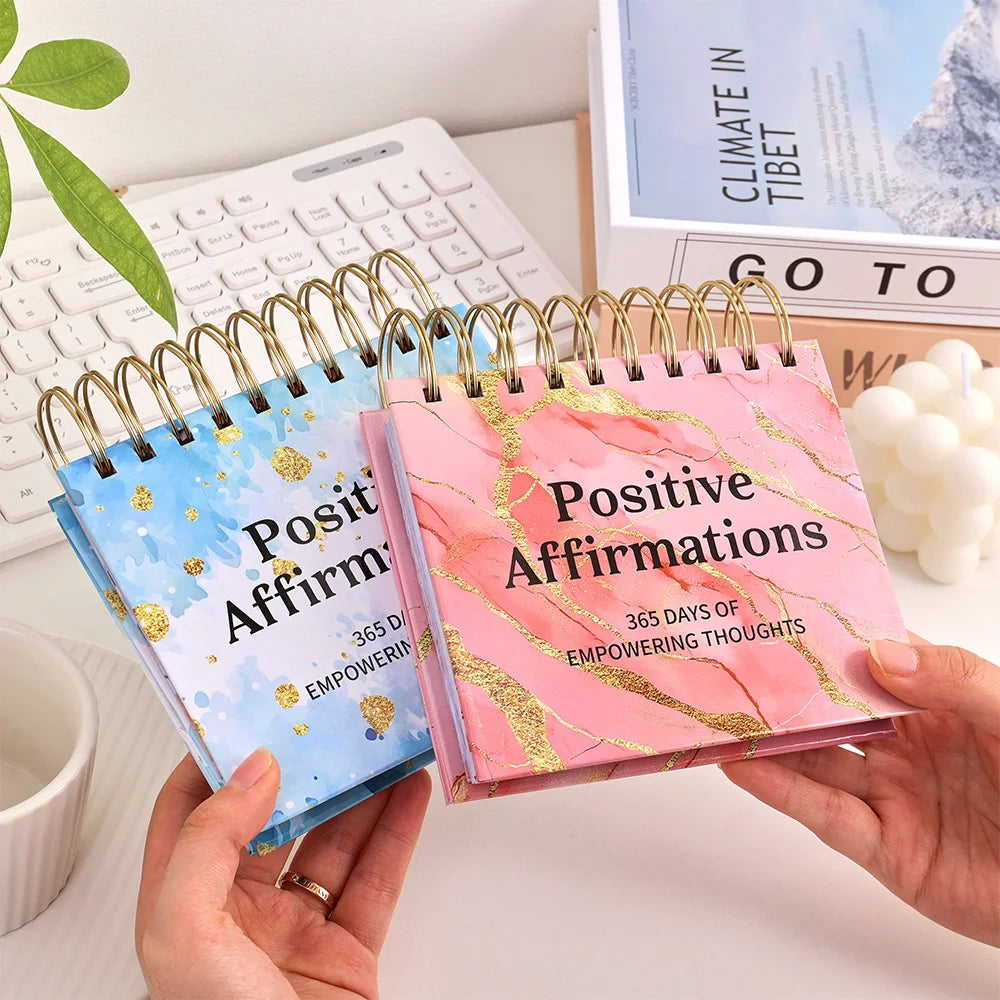 Two spiral-bound 'Positive Affirmations' notebooks held by hands on a desk.