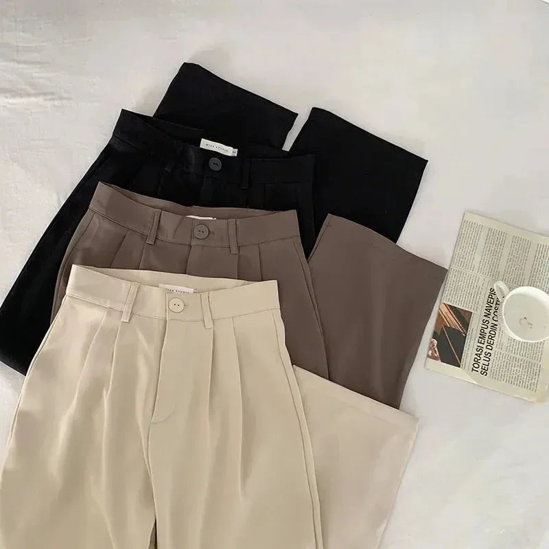 Three pairs of pants in black, brown, and beige on a light surface with a newspaper and cup.