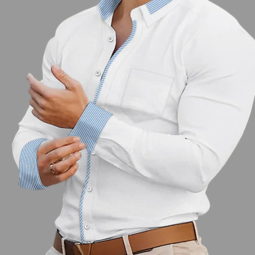 Man wearing a white shirt with blue striped cuffs and a brown belt, standing in an outdoor setting.