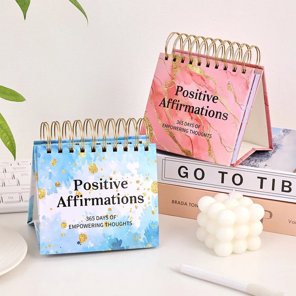 Two spiral-bound 'Positive Affirmations' calendars on a desk with a plant and books in the background.