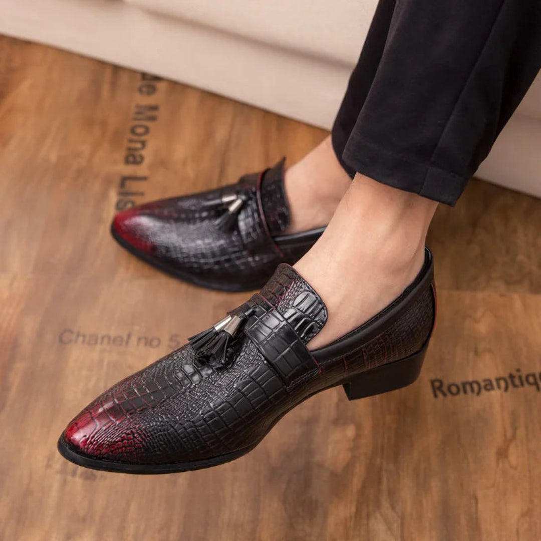 Black and red loafers with tassels worn by a person on a wooden floor.