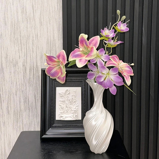 White vase with pink and purple flowers on a dark surface against a black frame and light gray wall.