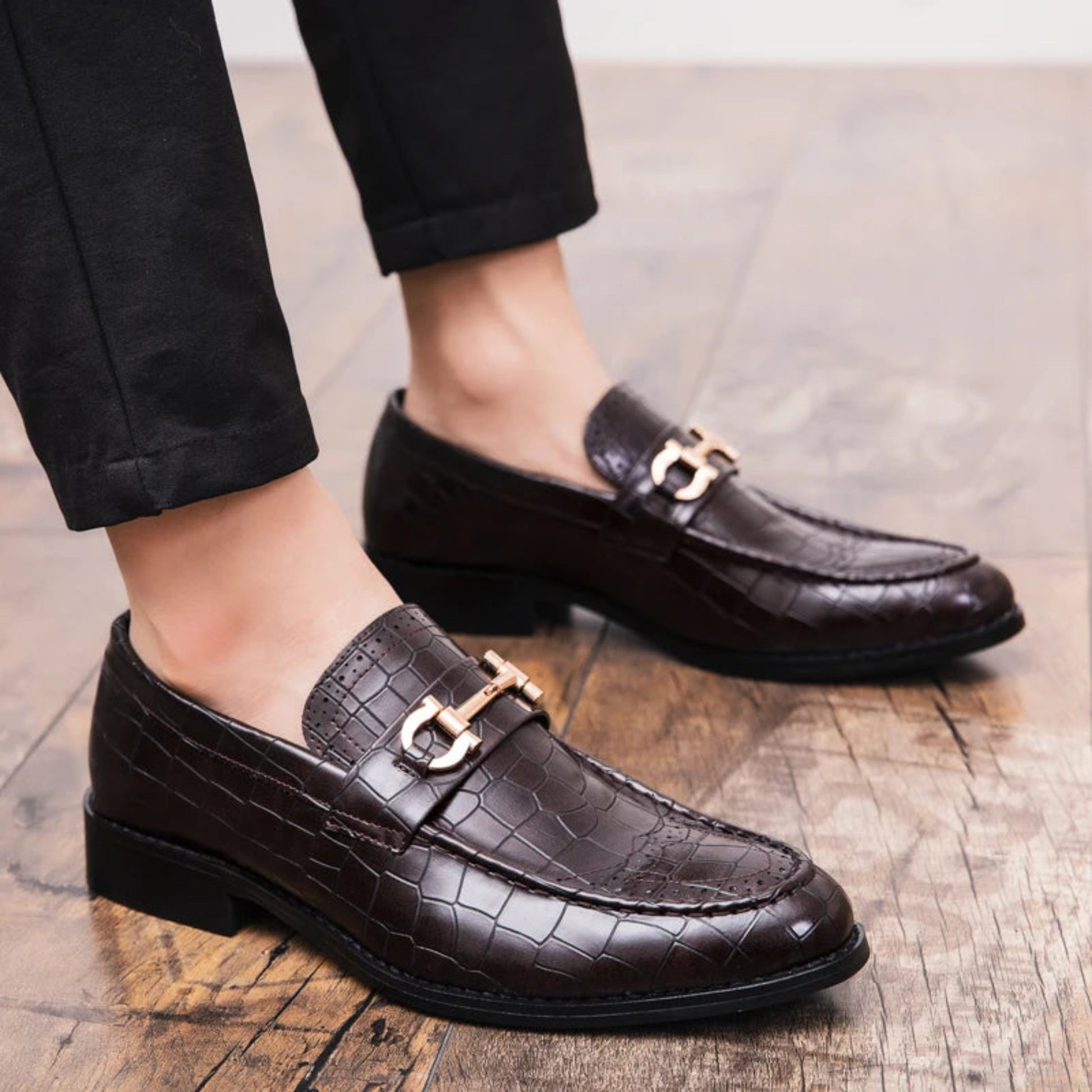Black leather loafers with gold buckles worn by a person on a wooden floor.