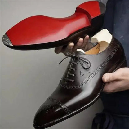 Brown leather dress shoe with red sole held by a hand against a neutral background