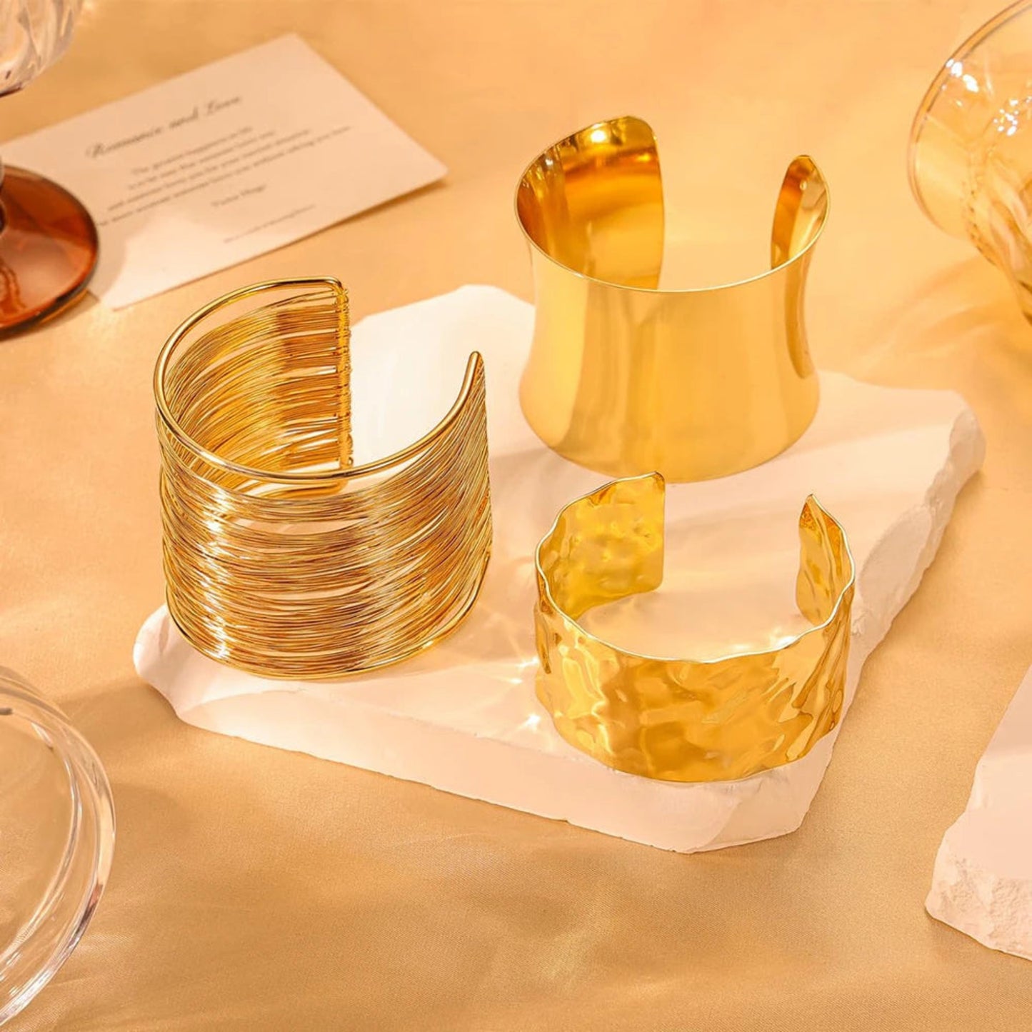 Three gold cuff bracelets on a white stone surface with a warm beige background.