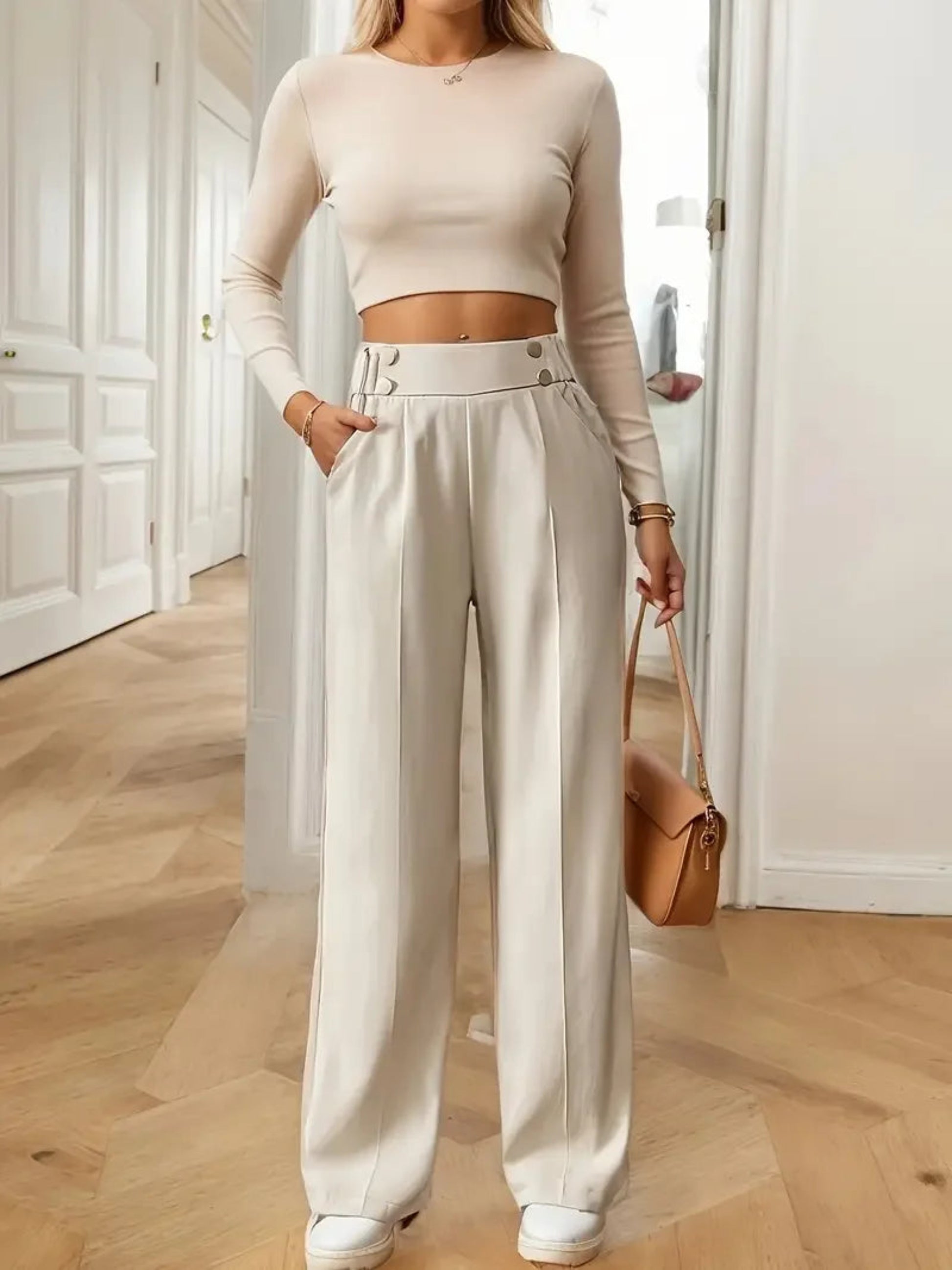 Person wearing a beige long-sleeve top and wide-leg pants, holding a brown handbag indoors.