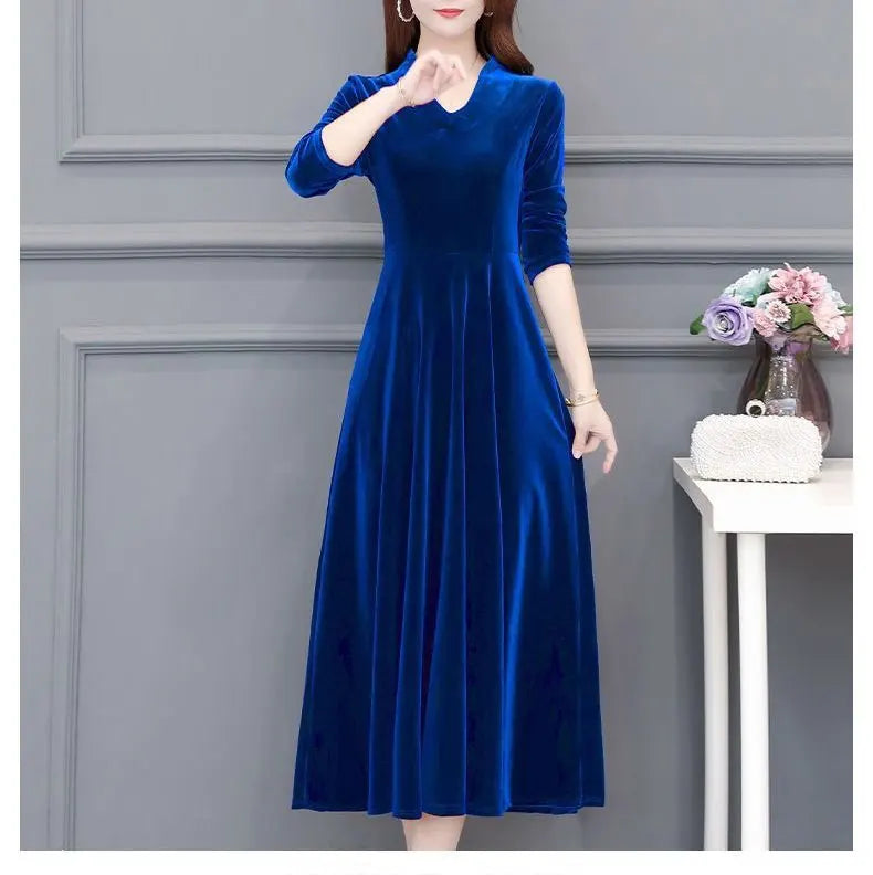 Woman wearing a blue dress against a gray wall with a small table and flowers in the background.