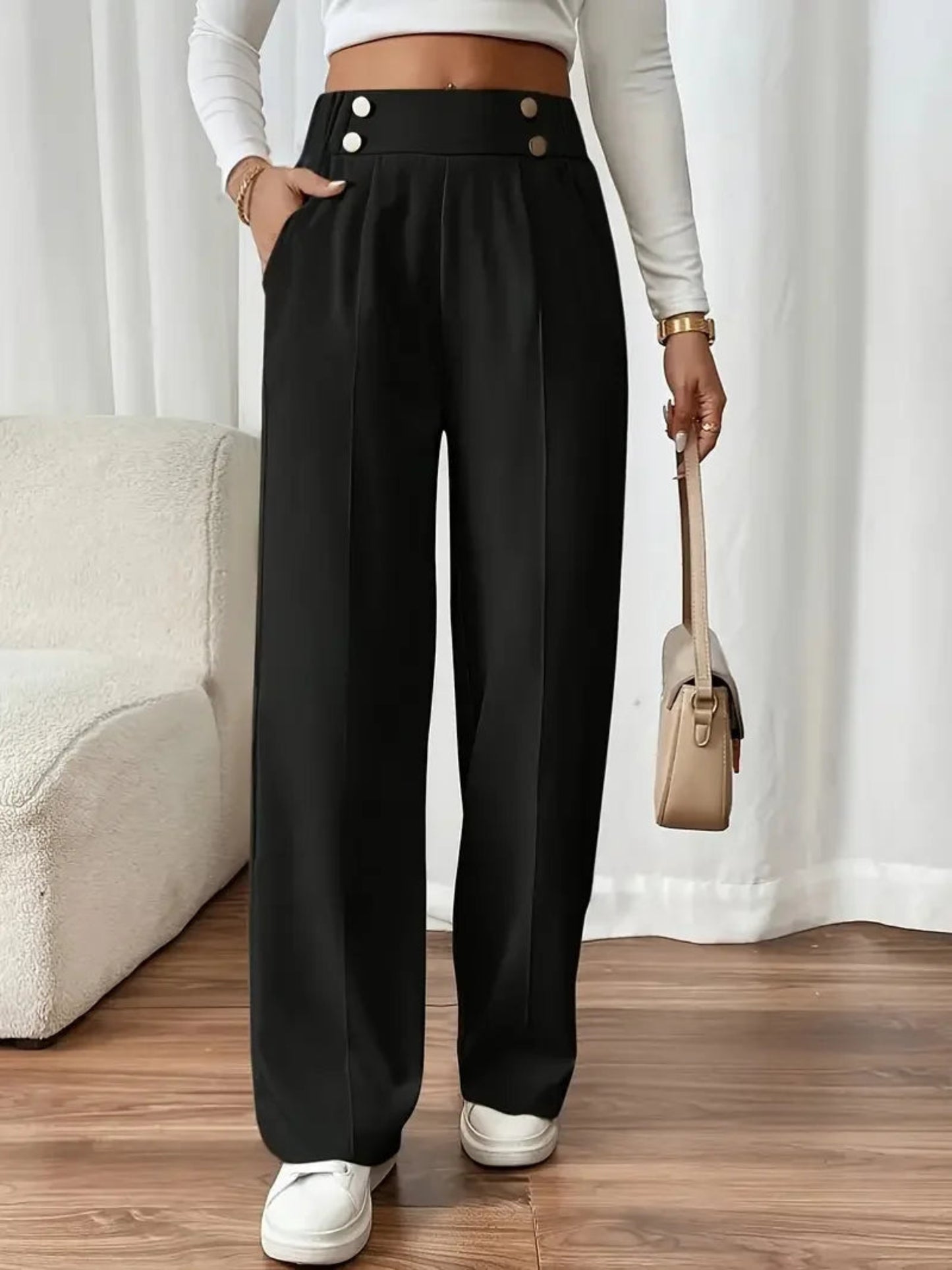 Person wearing black high-waisted pants with white buttons, standing in a room with a beige couch and white curtains.