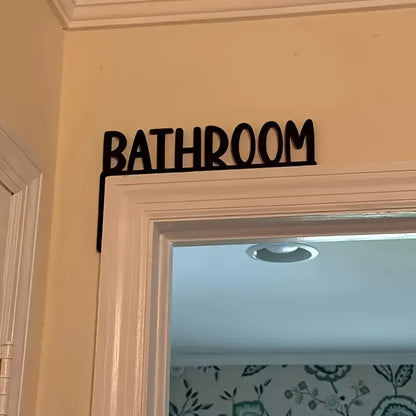 Black 'Bathroom' sign on a door frame with a light blue wall in the background.