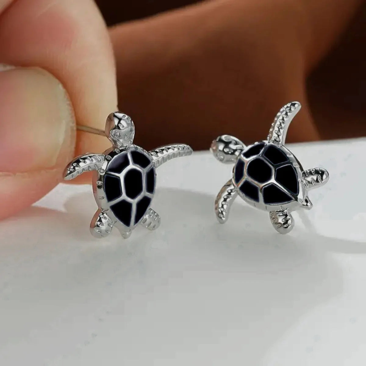 Turtle-shaped earrings with black details held between fingers on a white surface.