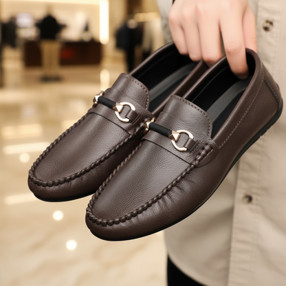 Brown loafers with gold buckles held by a hand against a neutral background